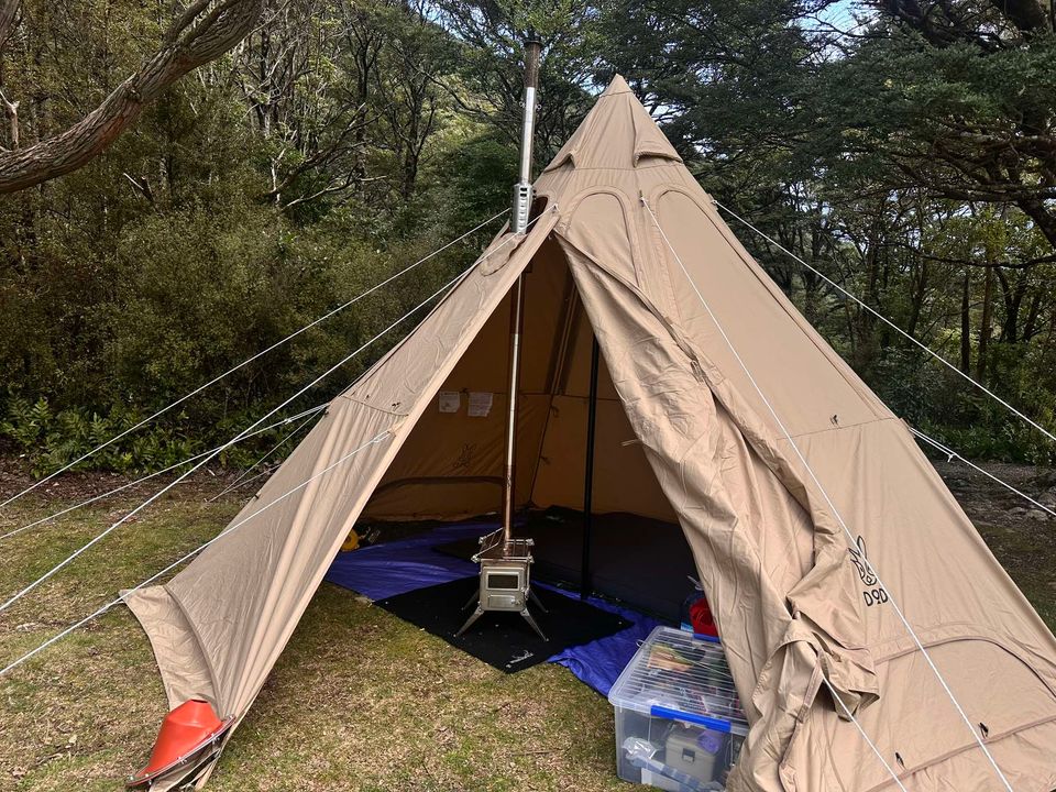 The DOD Outdoors Lotus Tipi | firetent Canvo™ is a spacious beige canvas tent set up on grass, partially open to reveal a wood stove and camping supplies by the entrance—ideal for winter camping among trees and bushes.