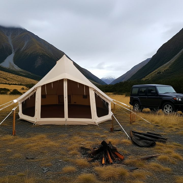 A Nomadic Canvascamp Apex 520 Premium Bell Tent stands on dry grass by a black SUV and campfire, with tall mountains and a cloudy sky in the background—perfect for professional use or outdoor adventure.