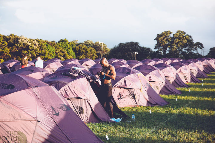 A shirtless man stands beside a row of Nomadic North Face Oval Intention | Event Collaboration tents, set up on a grassy field surrounded by trees under partly cloudy skies. The used tents are evenly spaced and the campsite looks organized.