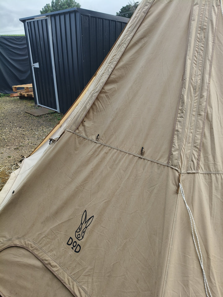 A used Nomadic DoD Lotus Firetent 4m in tan, featuring the DOD rabbit logo, is set up outdoors near a black metal shed and stacked pallets, similar to surplus inventory.