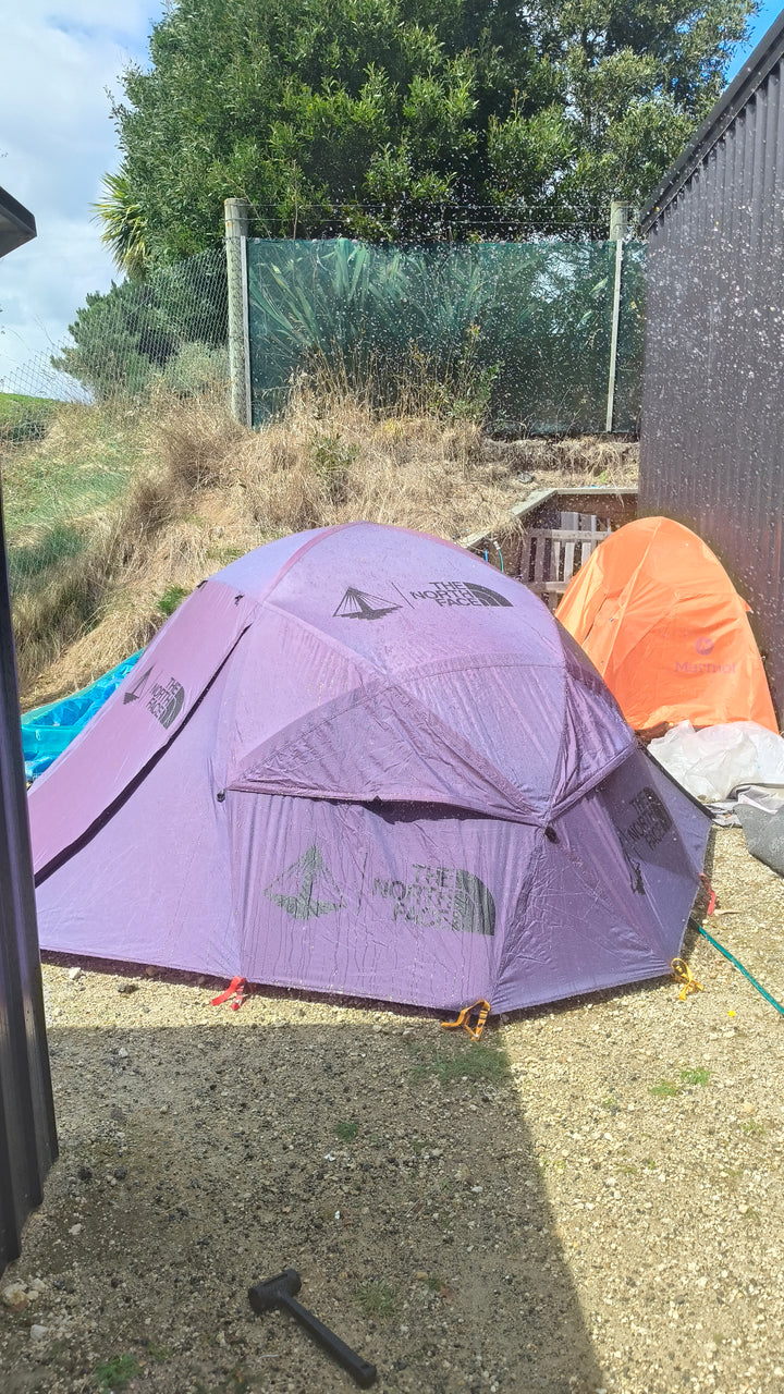 Two Nomadic North Face Oval Intention | Event Collaboration tents—one purple and one orange—are pitched on grass near a black fence and wooden building, as water sprays over them. Trees and a wire fence are visible in the background.
